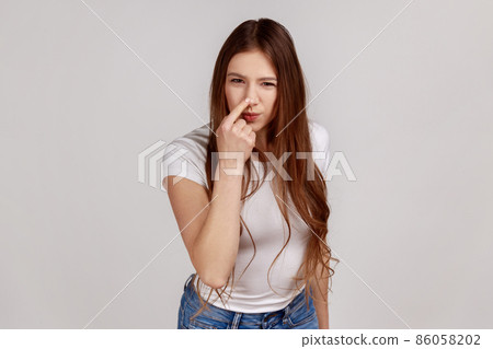 That's lie, you cheat. Disgruntled woman looking angrily at camera and showing lie gesture, touching nose meaning falsehood, wearing white T-shirt. Indoor studio shot isolated on gray background. 86058202