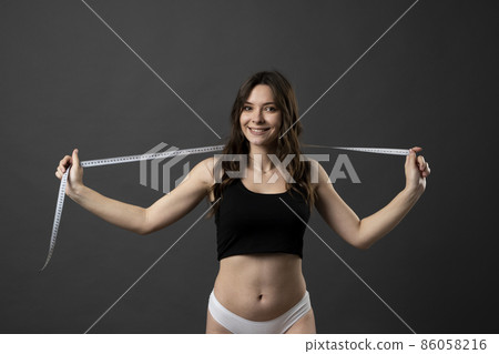 Woman in a black top and white panties with a white measuring tape, in a black background. Healthy lifestyle concept. Diet. 86058216