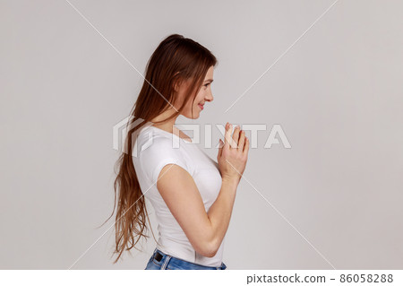 Side view portrait of scheming devious woman clasping hands and thinking over tricky plan, having sly cunning idea to prank, wearing white T-shirt. Indoor studio shot isolated on gray background. Side view portrait of scheming devious woman clasping hands and thinking over tricky plan, having sly cunning idea to prank, wearing white T-shirt. Indoor studio shot isolated on gray background. 86058288