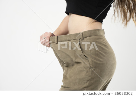 Woman showing result after weight loss wearing on old jeans on gray background. Diet concept and weight loss. Woman showing result after weight loss wearing on old jeans on gray background. Diet concept and weight loss. 86058302
