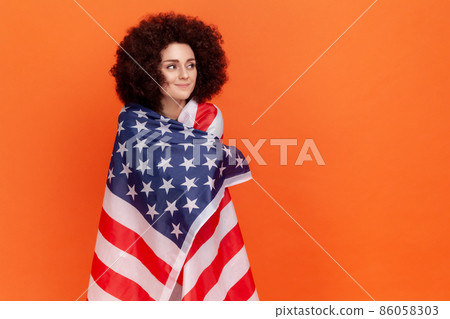 Calm dreamy woman with Afro hairstyle wearing green casual style sweater standing wrapped in USA flag, looking away, human rights. Indoor studio shot isolated on orange background. Calm dreamy woman with Afro hairstyle wearing green casual style sweater standing wrapped in USA flag, looking away, human rights. Indoor studio shot isolated on orange background. 86058303