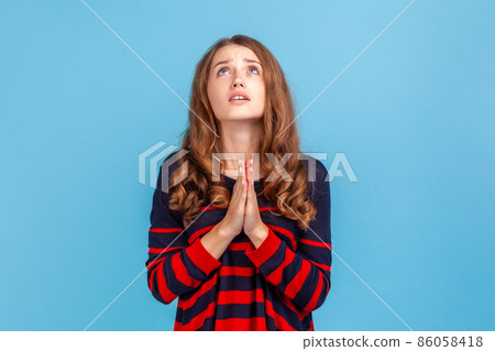 Portrait of young attractive woman wearing striped casual style sweater looking up with imploring desperate grimace, praying to god asking for help. Indoor studio shot isolated on blue background. Portrait of young attractive woman wearing striped casual style sweater looking up with imploring desperate grimace, praying to god asking for help. Indoor studio shot isolated on blue background. 86058418