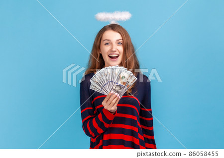 Shocked angelic woman wearing striped casual style sweater and nimb over head, holding fan of dollars banknotes and looking at camera with open mouth. Indoor studio shot isolated on blue background. 86058455