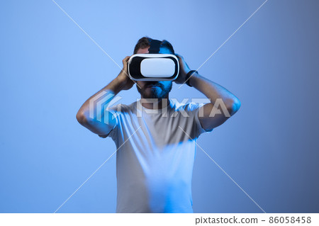Young bearded guy in VR goggles touching air and excited with new feelings in dark neon lights. 86058458