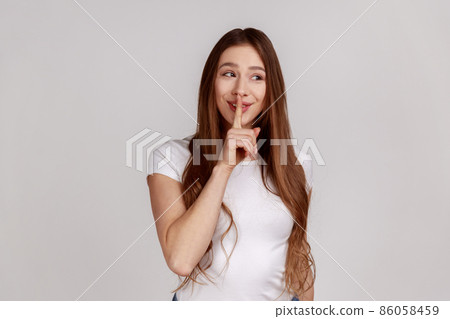 Shh, it's big secret. Beautiful cunning woman smiling, showing gesture secret sign with finger near her lips, wearing white T-shirt. Indoor studio shot isolated on gray background. 86058459