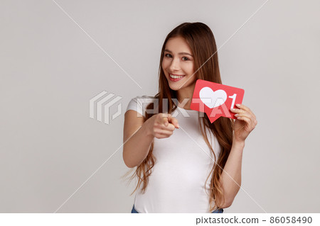 Portrait of dark haired positive woman blogger holding hear like icon and pointing finger at camera, wearing white casual style T-shirt. Indoor studio shot isolated on gray background. 86058490