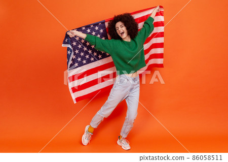 Full length portrait of happy woman with Afro hairstyle wearing green casual style pretending flying, having fun, holding american flag. Indoor studio shot isolated on orange background. Full length portrait of happy woman with Afro hairstyle wearing green casual style pretending flying, having fun, holding american flag. Indoor studio shot isolated on orange background. 86058511