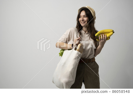 Zero waste concept. Young brunette woman holding cotton eco bag with organic fruits and vegetables on a shoulder and holding bananas in a hand. Using reusable crochet net bag for grocery shopping. 86058548