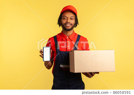 Pleased friendly courier in red T-shirt and blue overalls holding cardboard box and smart phone with empty display for delivery service adverisement. Indoor studio shot isolated on yellow background. 86058591