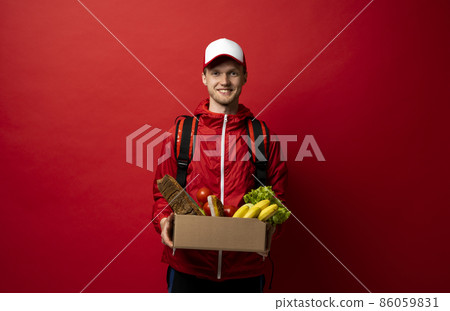 Delivery man in red uniform holds cardboard paper box with food isolated on white background, studio portrait. Service concept. 86059831