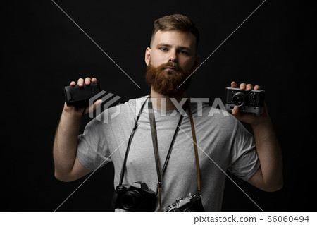 Professional photographer in a grey t-shirt with a bunch of different cameras in a hands and on a shoulder looking on a camera and ready for make a good shoot on a black background. Professional photographer in a grey t-shirt with a bunch of different cameras in a hands and on a shoulder looking on a camera and ready for make a good shoot on a black background. 86060494
