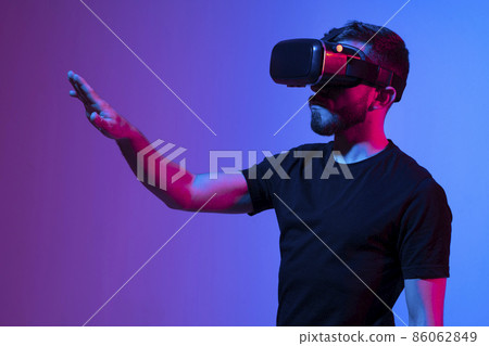 Portrait of man using VR headset while communicating with a friends in a metaverse in dark interior illuminated neon light. Close-up futuristic googles with colored light. Portrait of man using VR headset while communicating with a friends in a metaverse in dark interior illuminated neon light. Close-up futuristic googles with colored light. 86062849