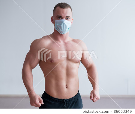 Male bodybuilder in a medical mask with muscular bare chest. The guy goes in for sports in quarantine. Respiratory protection. 86063349