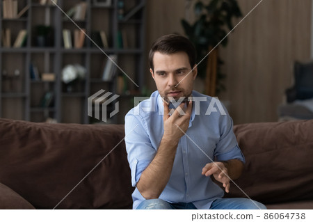 Serious man recording audio message on smartphone, sitting on couch Serious man recording audio message on smartphone, sitting on couch 86064738