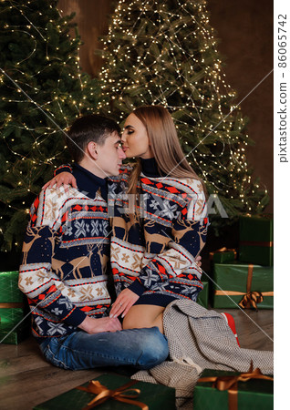 A guy and a girl hug and kiss against the background of a Christmas tree and a red sofa A guy and a girl hug and kiss against the background of a Christmas tree and a red sofa 86065742