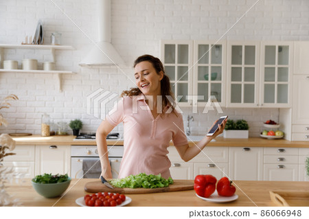 Happy carefree young woman dancing to music, cooking in kitchen. 86066948