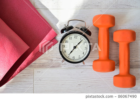 Top view on orange dumbbell alarm clock and raspberry yoga mat. The concept of home sports during quarantine. Fitness time. Early rise for classes. Top view on orange dumbbell alarm clock and raspberry yoga mat. The concept of home sports during quarantine. Fitness time. Early rise for classes. 86067490