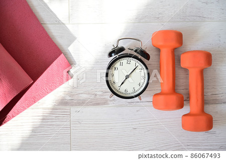 Top view on orange dumbbell alarm clock and raspberry yoga mat. The concept of home sports during quarantine. Fitness time. Early rise for classes. 86067493