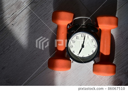 It's time to do sports early in the morning. Top view on alarm clock and orange dumbbells on a wooden floor. 86067510