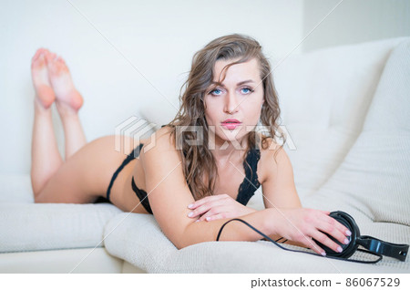 Beautiful woman in black lace underwear lies on her stomach and listens to music. The girl in the bedroom relaxes the bed holds headphones in her hands. Beautiful woman in black lace underwear lies on her stomach and listens to music. The girl in the bedroom relaxes the bed holds headphones in her hands. 86067529