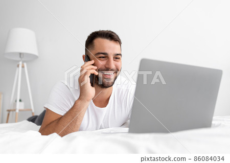 Happy young attractive guy with beard lies on white bed call by phone, looks at laptop, working or chatting Happy young attractive guy with beard lies on white bed call by phone, looks at laptop, working or chatting 86084034