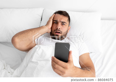 Shocked upset millennial attractive guy with beard reads message on phone and presses his hand to face Shocked upset millennial attractive guy with beard reads message on phone and presses his hand to face 86084075