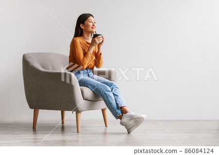 Beautiful young Asian woman drinking coffee or tea, having break, relaxing in armchair against white studio wall Beautiful young Asian woman drinking coffee or tea, having break, relaxing in armchair against white studio wall 86084124