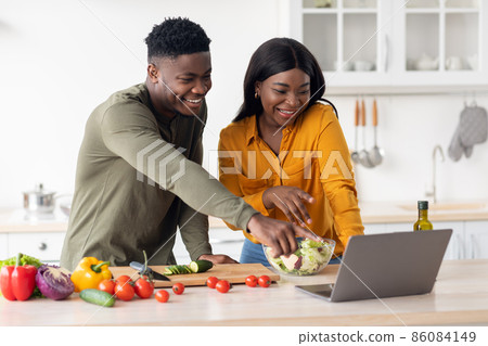 Cheerful Black Young Couple Using Laptop While Cooking Healthy Food In Kitchen Cheerful Black Young Couple Using Laptop While Cooking Healthy Food In Kitchen 86084149