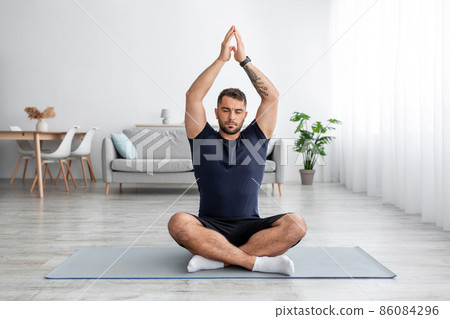 Calm young handsome guy blogger with beard with closed eyes practices yoga at home in living room Calm young handsome guy blogger with beard with closed eyes practices yoga at home in living room 86084296