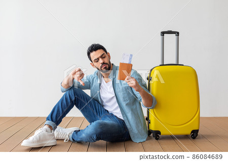 Bad travel agency service. Young arab man holding passport with tickets, showing dislike gesture, sitting near suitcase 86084689