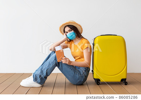 Upset woman wearing protective medical mask, holding passport with ticket and boarding pass, sitting near her luggage Upset woman wearing protective medical mask, holding passport with ticket and boarding pass, sitting near her luggage 86085286
