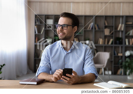 Smiling dreamy businessman wearing glasses looking in distance, holding smartphone 86085305