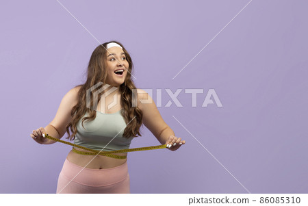 Happy excited caucasian plus size woman in sportswear measures her waist with measuring tape and enjoy result 86085310