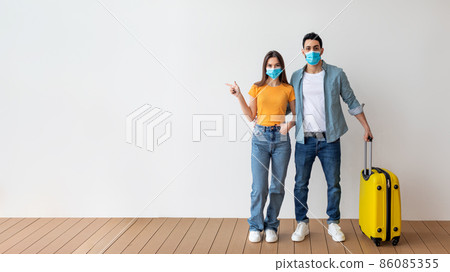 Young spouses wearing medical face masks standing with suitcase, woman pointing aside at free space Young spouses wearing medical face masks standing with suitcase, woman pointing aside at free space 86085355