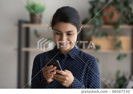 Happy Indian girl smiling at smartphone screen, holding mobile phone Happy Indian girl smiling at smartphone screen, holding mobile phone 86087530