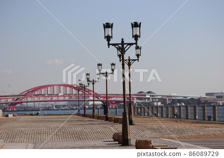 Cobblestone coastal park with street lights, blue sea, red bridge where cars come and go, ship, harbor, building cityscape, blue sky and cloud view 86089729