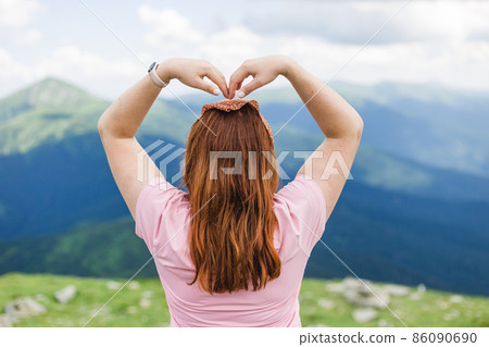 Young woman holding her hands up in the shape of heart against the background of the mountain valley. Happy girl standing backwards looking away. Traveling in the mountains. Freedom, travel concept Young woman holding her hands up in the shape of heart against the background of the mountain valley. Happy girl standing backwards looking away. Traveling in the mountains. Freedom, travel concept 86090690