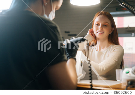 Close-up view from back of manicure master removing gel polish or acrylic from nails with help of manicure machine. Happy smiling young woman getting nail treatment at beauty salon at table with lamp. 86092600