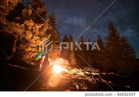 Night camping near forest. Rear view couple of tourists spending romantic evening outdoors by bonfire on lawn. On the background tents, pines, starry sky. Active recreation in nature. Night camping near forest. Rear view couple of tourists spending romantic evening outdoors by bonfire on lawn. On the background tents, pines, starry sky. Active recreation in nature. 86092897