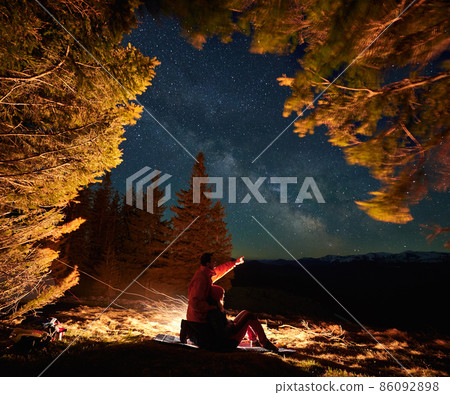 Side view of romantic couple in the mountains resting by campfire on tourist rug among meadow under starry sky. Guy hugging his girlfriend by her back and pointing her to magical Milky Way galaxy. 86092898