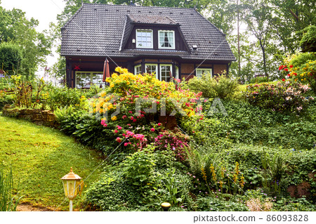 Well-kept garden with flowering shrubs and bushes in front of large luxury family house with garden light in foreground Well-kept garden with flowering shrubs and bushes in front of large luxury family house with garden light in foreground 86093828