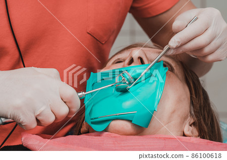 A patient with a cheek retractor with a green cofferdam in a dental clinic, a dentist in latex gloves examines her teeth with a boron and a mirror. A patient with a cheek retractor with a green cofferdam in a dental clinic, a dentist in latex gloves examines her teeth with a boron and a mirror. 86100618