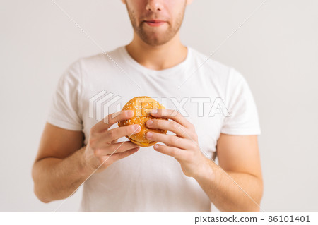Close-up cropped shot of unrecognizable hunger young man holding in hands delicious burger on white isolated background. Studio shot of happy handsome male eating tasty meal. 86101401