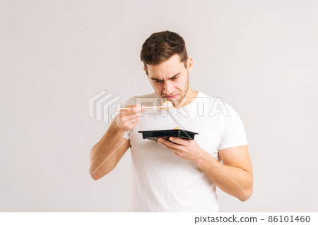 Portrait of disgusted young man holding sushi with chopsticks and sniffs with look of revulsion on face on white isolated background. Sad Caucasian guy dissatisfied with bad quality Asian food. 86101460