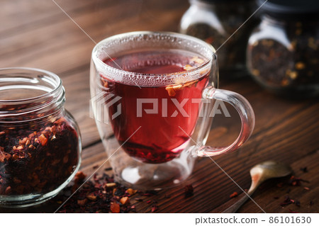 Glass cup of hot aromatic red fruit tea, jar of natural fruit tea with hibiscus petals, fruit slices and berries. Jars of aromatic fruit tea on background. Glass cup of hot aromatic red fruit tea, jar of natural fruit tea with hibiscus petals, fruit slices and berries. Jars of aromatic fruit tea on background. 86101630