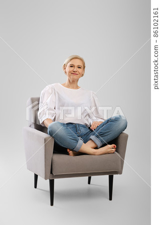 Barefoot senior woman sitting in an armchair crossing legs in studio 86102161