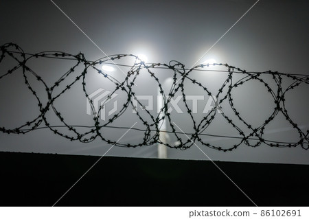 silhouette of barbed wire on a fence against a background of blurry lamp posts at foggy night 86102691