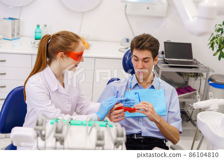 Ginger woman in white robe smiling and giving protective goggles to young man during appointment in modern dental clinic 86104810