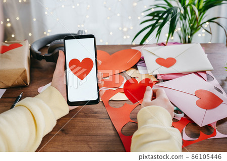 Woman holding a smartphone while making a Valentine greeting cards Woman holding a smartphone while making a Valentine greeting cards 86105446