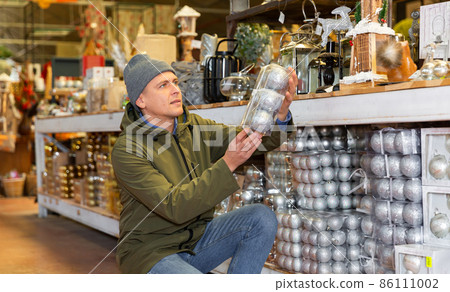 Preparing for the New Year - happy man chooses Christmas tree decorations in the warehouse of store 86111002